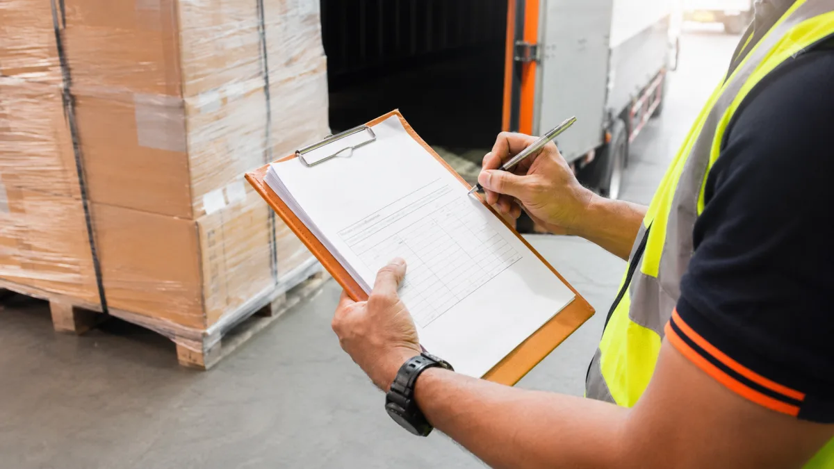 Warehouse worker checking and signing a consignment note beside a pallet being loaded for freight